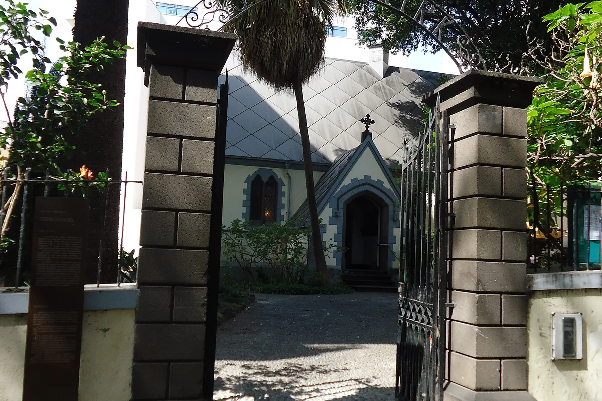 An open iron gate between two stone pillars, with the roof of a small church visible behind palms in a garden