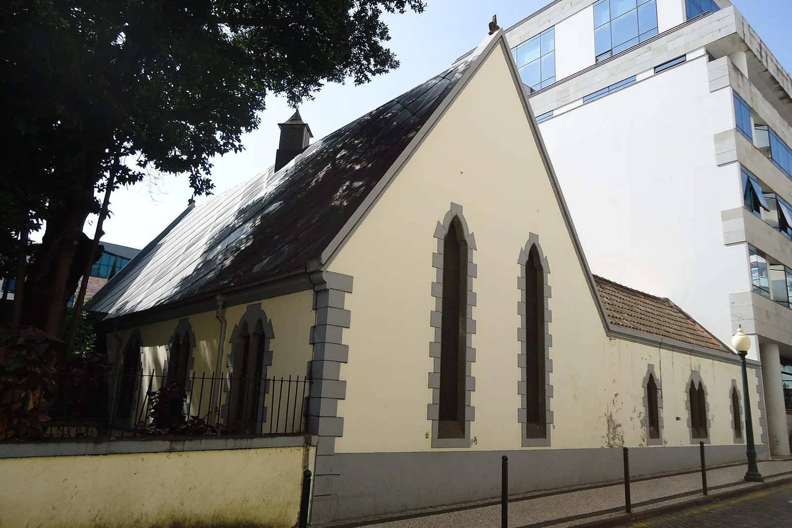 The long side wall of the church seen from the street, with pointed windows, a gabled roof and a modern building rising behind